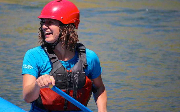 a person wearing a life jacket and helmet smiles while sitting in a raft and using a paddle